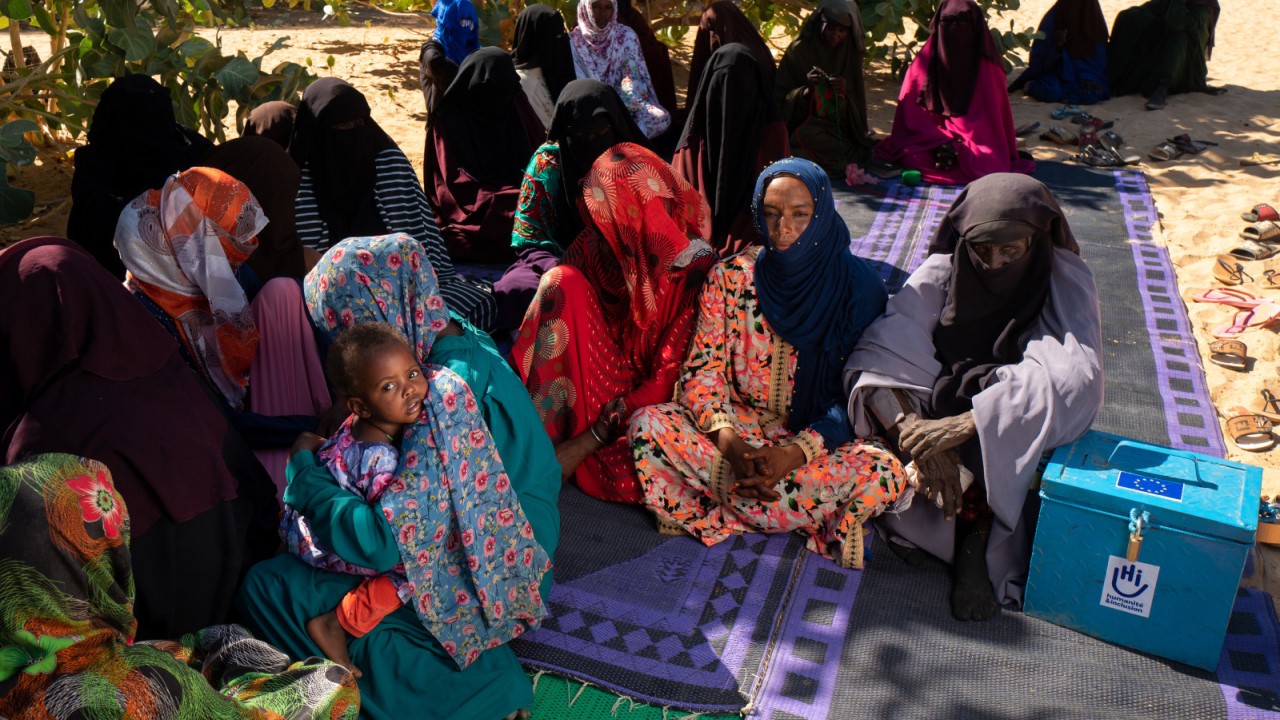 Plusieurs femmes sont assises sur des tapis installés sur le sable, elles attendent à l'ombre des arbres.