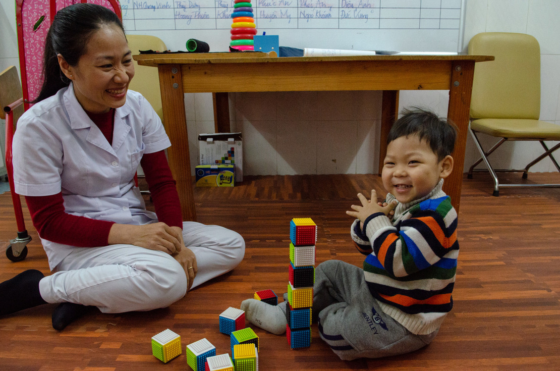 Un petit garçon applaudit à coté de la tour qu'il a construite avec des cubes devant l'ergothérapeuthe de HI