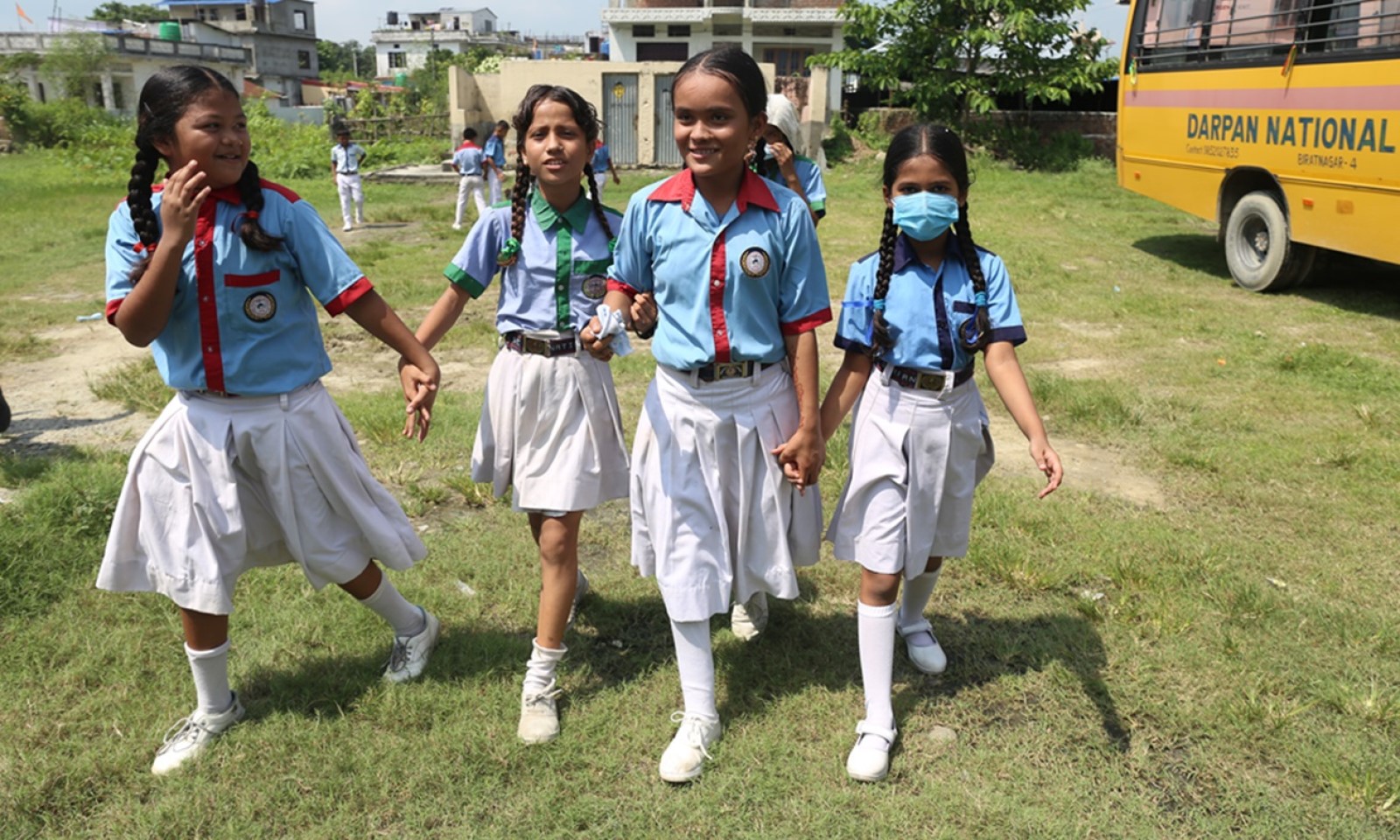 Four girls from Nepal in school uniform are holding hands while walking outside. One has a prothesis. In the back, there is a school bus. 