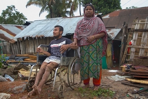 In front of houses destroyed by floods, a man in a wheelchair and a woman at his side.