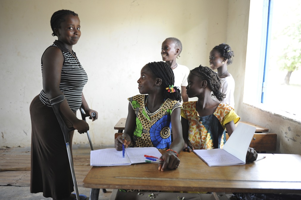 A classroom setting with four girls from Senegal sitting at a wooden desk, two of whom are writing in notebooks. An adolescent girls of 14 years old with crutches stands beside them, looking directly at the camera with a smile.