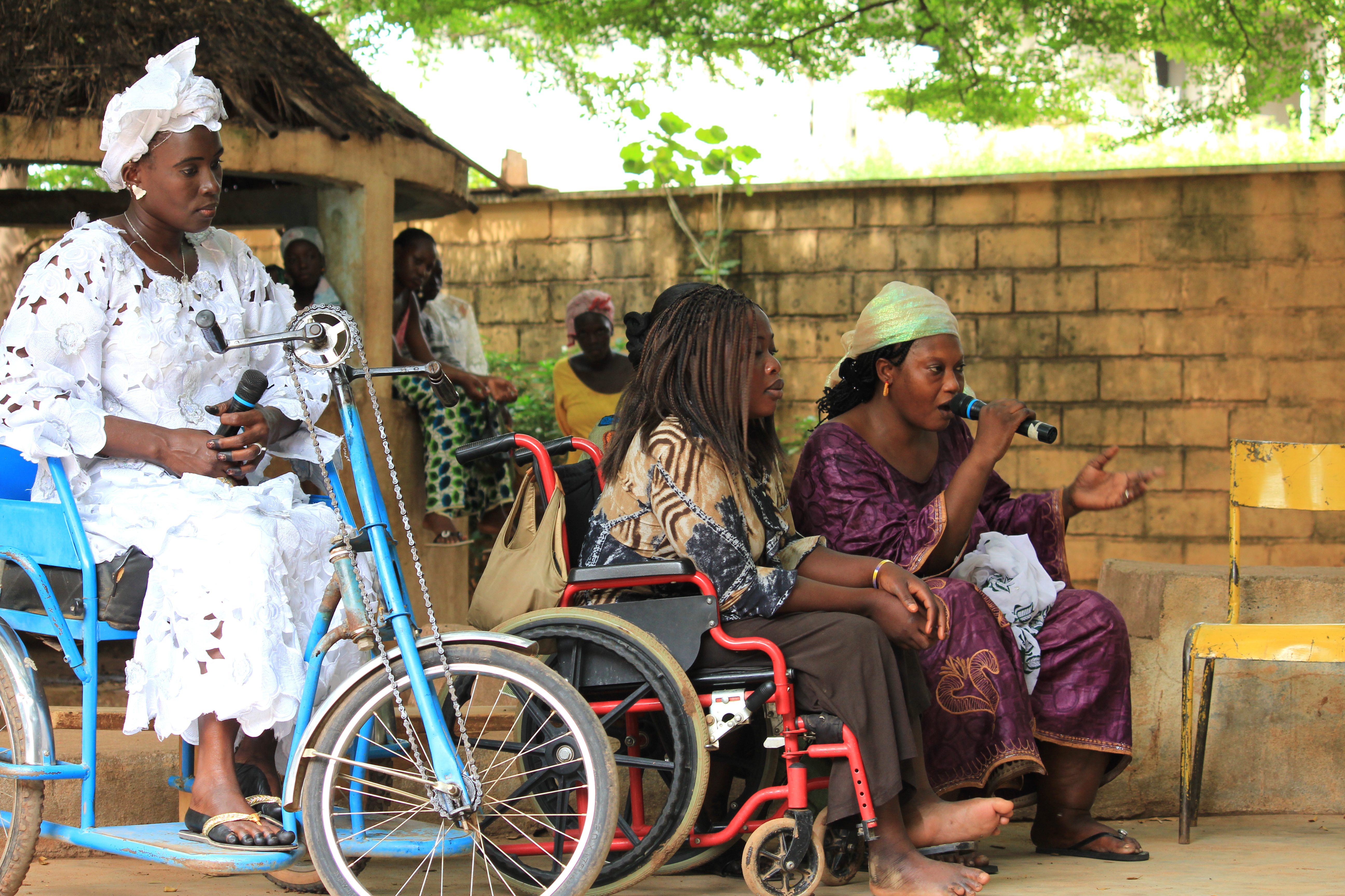 Three seated women, two of them in wheelchairs, speak into a microphone in a village square.; }}