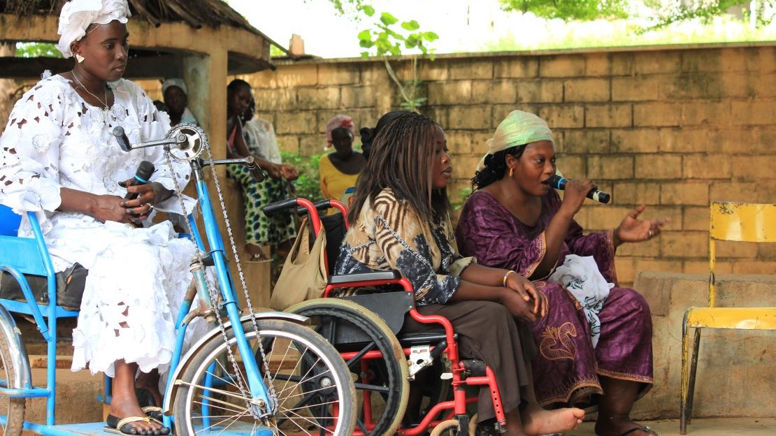 Three seated women, two of them in wheelchairs, speak into a microphone in a village square.; }}