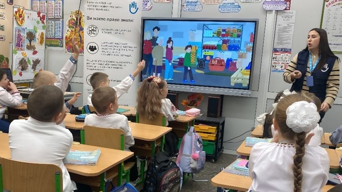 Des enfants sont assis à leurs bureaux dans une classe face à un écran où l'on montre des images pédagogiques, une famille ukrainienne dans sa vie de tous les jours pour expliquer les comportements sûrs à adopter en cas de danger, au tableau l'agent de sensibilisation de HI se tient debout et leur décrit les images.; }}