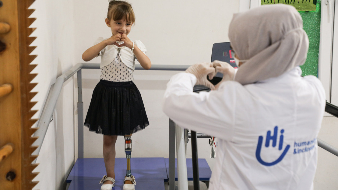 Photo d'Enas, une petite fille de 5 ans qui porte une jupe noire avec un haut et un gilet blanc. Elle est debout sur un escalier de rééducation avec des rampes. On voit ses jambes, dont sa prothèse de jambe. Enas regarde la travailleuse HI en face d'elle. Enas et Fatima, la travailleuse HI, font un cœur avec leurs mains.; }}