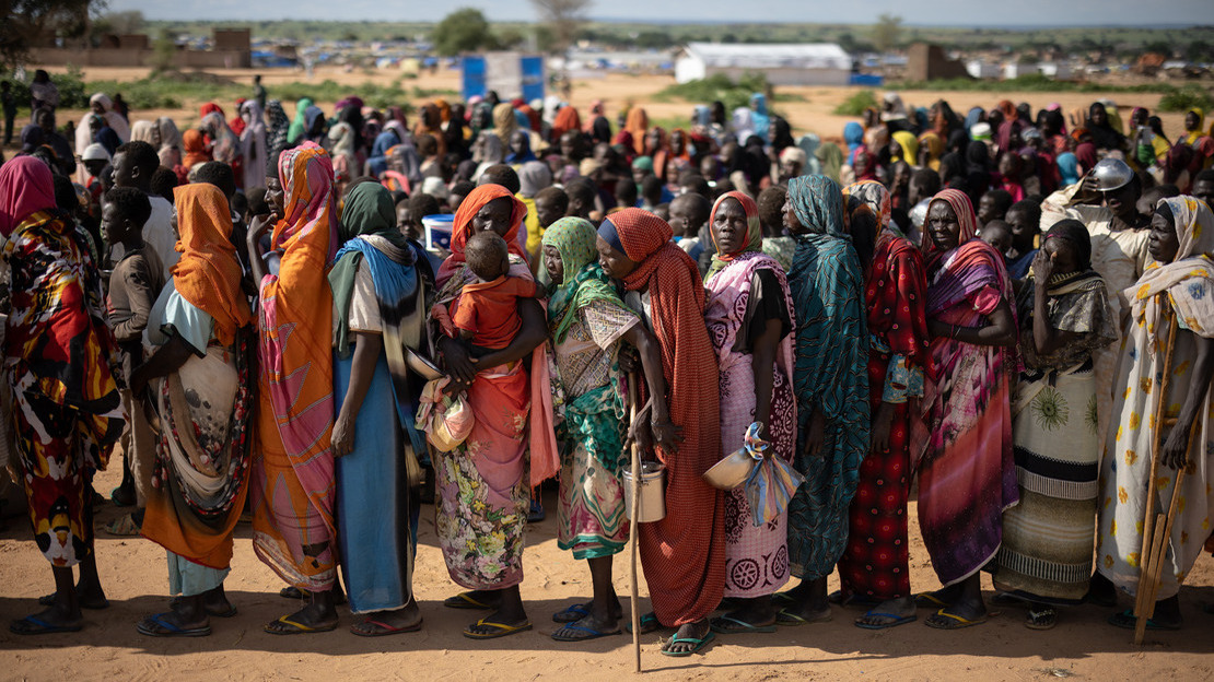 Women stand one behind the other, in a line that can be seen continuing far behind them. Each is holding an empty basin or container.; }}