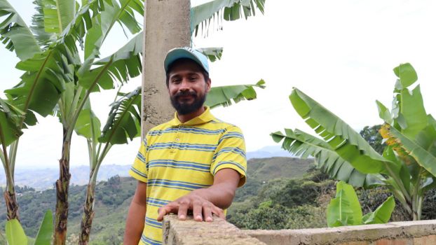 Carlos Bolaños at his home in the Cauca department. Portrait of a man leaning on a low wall and smiling at the camera. Behind him, we see banana tree branches and, in the distance, green hills.; }}