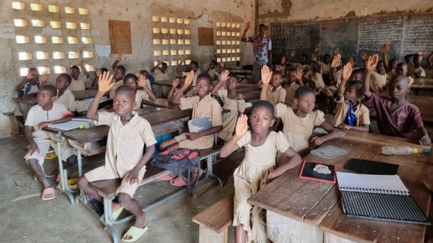 Year 2 class taught by Yaovi Ahli at Kombonloaga public primary school in Dapaong, northern Togo. Inside a classroom. Children aged 8–10 in beige uniforms are seated around wooden desks and raising their hands.; }}