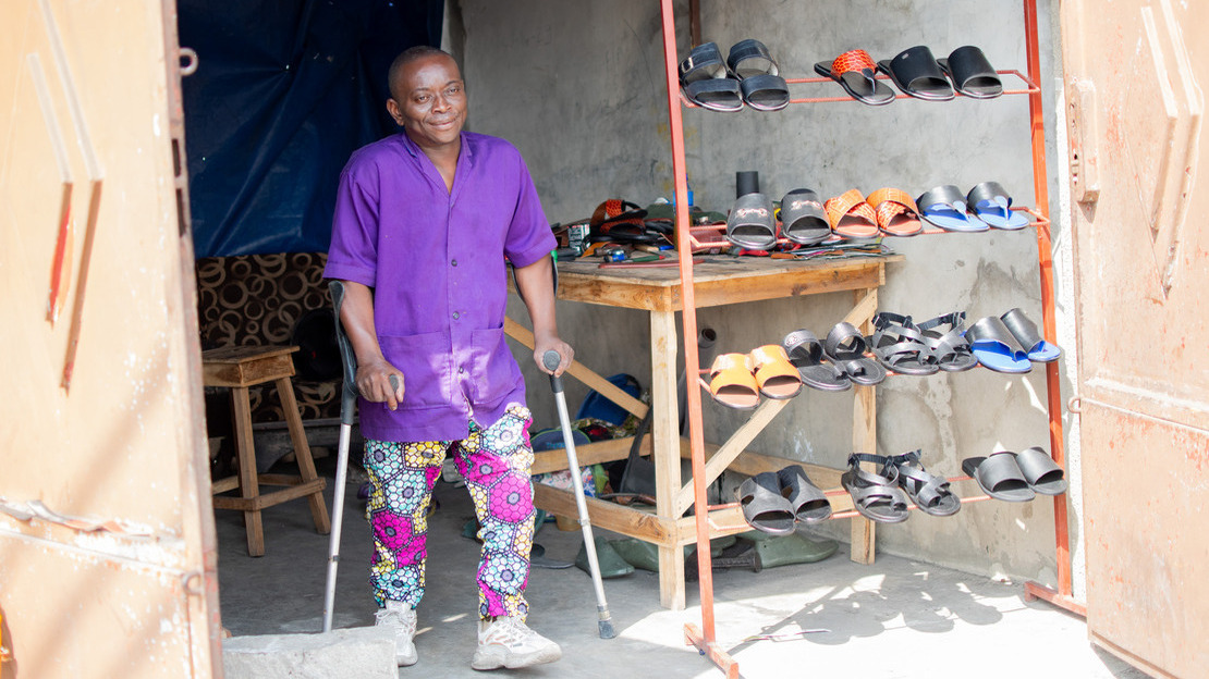 A man stands on crutches between the open doors of a workshop overlooking the street. A display case shows different pairs of sandal-type shoes.; }}