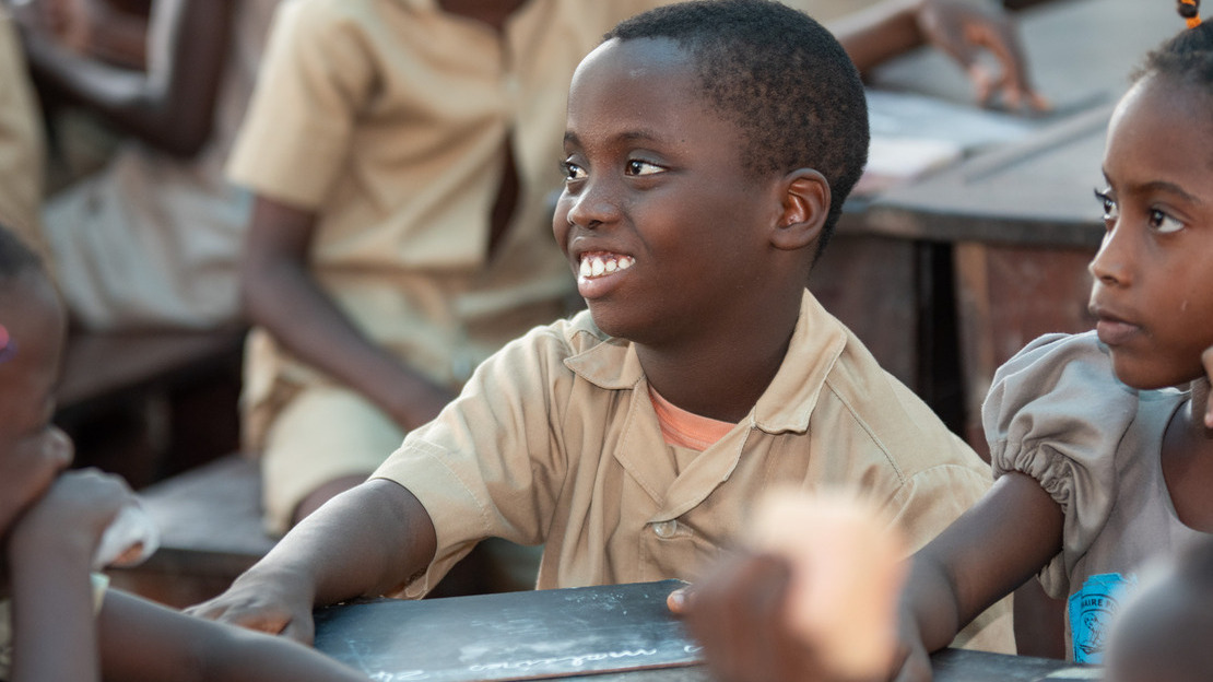 Close-up portrait of a boy sitting in a classroom, holding a slate in his hand. His face is turned three-quarters and he is smiling. Around him, we can make out the faces and arms of other children.; }}