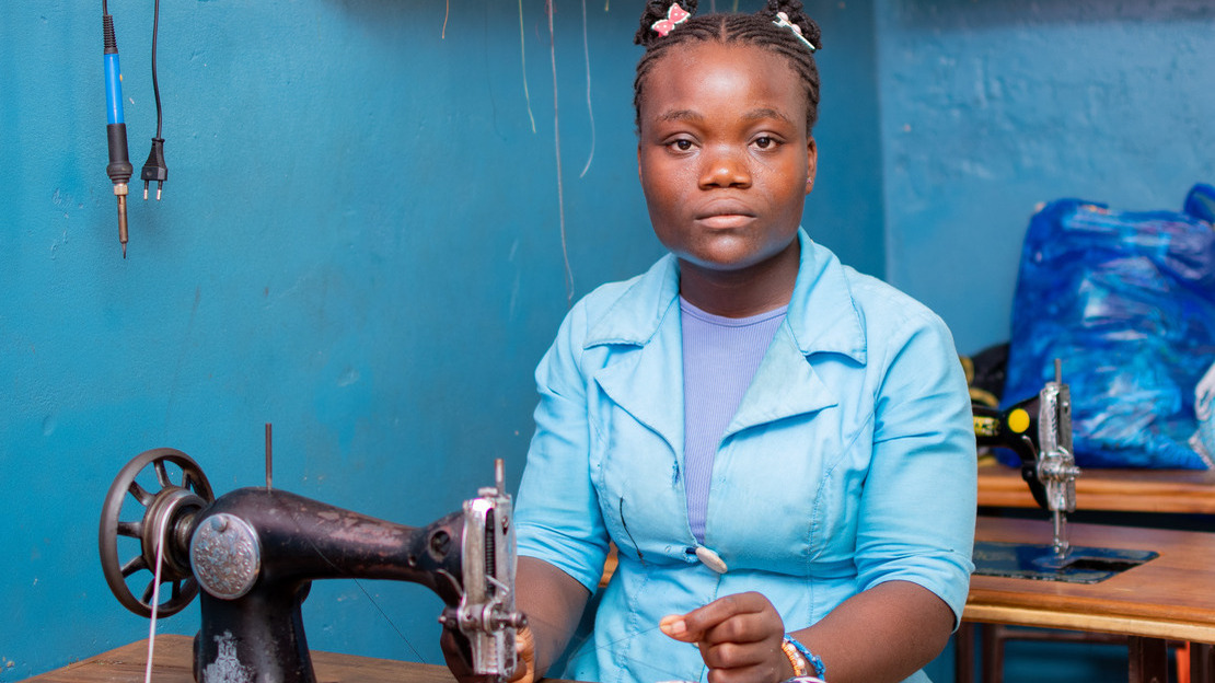 Portrait of Belvine seated at a table, looking at the camera. In front of her is a sewing machine and a piece of fabric. She pulls a thread through the machine.; }}