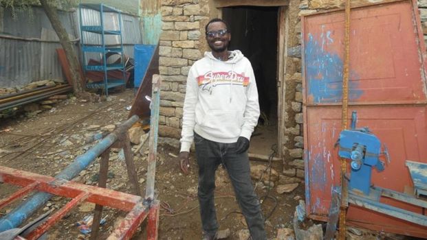 A young men stand in front of is workshop.; }}