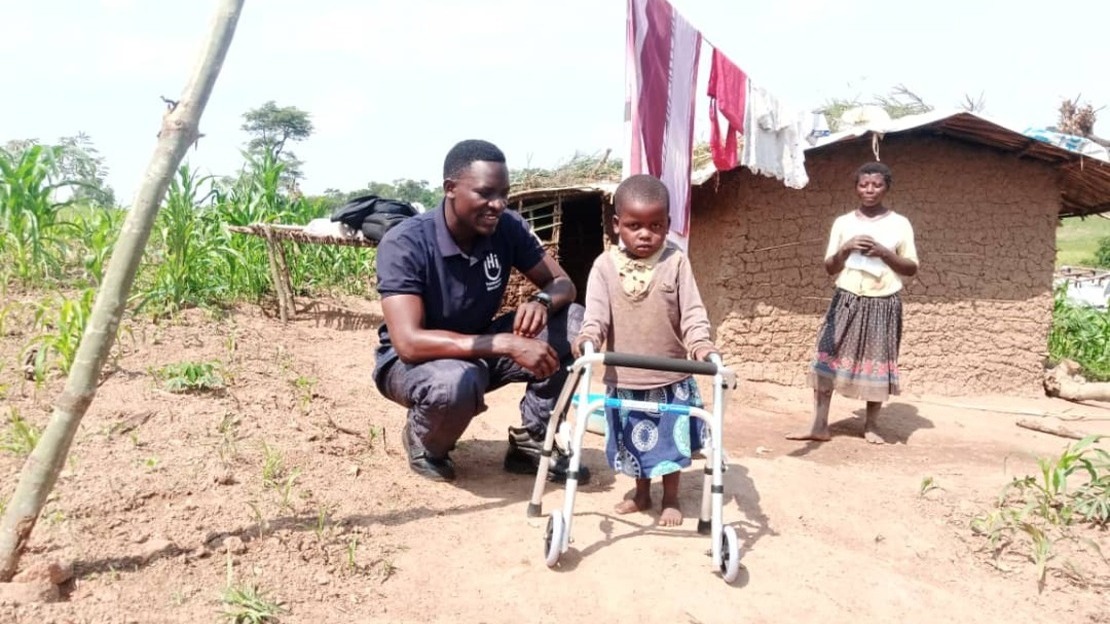 In front of a brick house, a little girl stands leaning on a walking frame. Kneeling beside her is a smiling man. A woman stands in the background.; }}