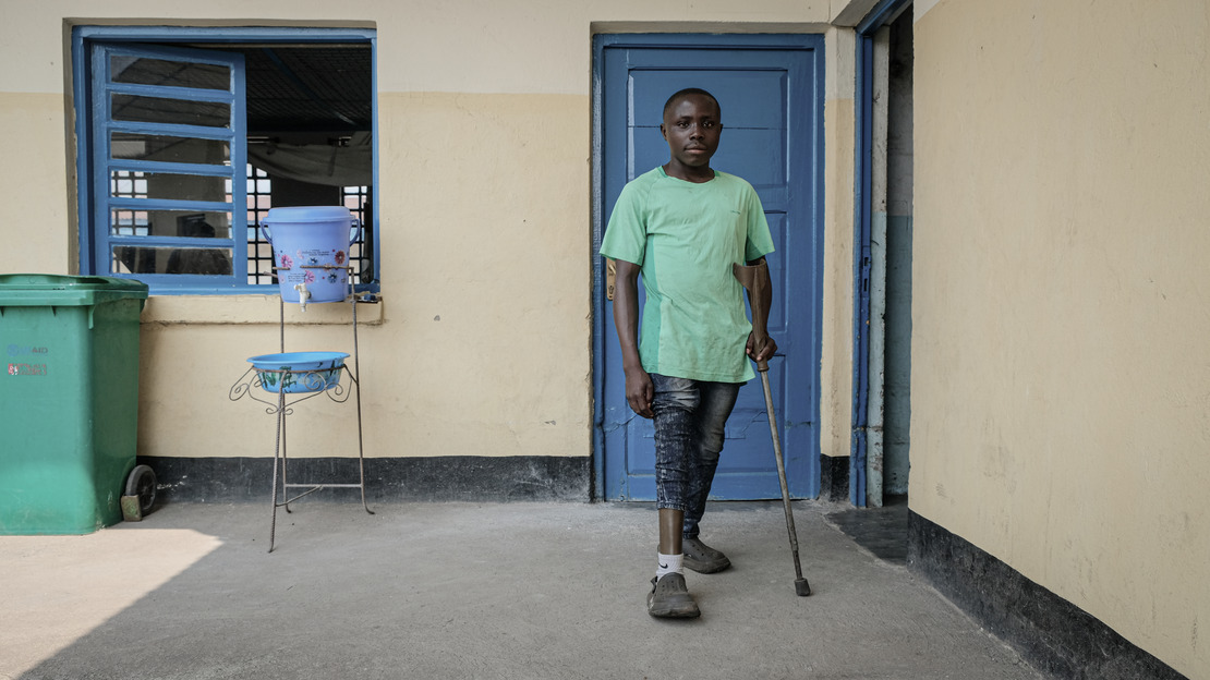 Un homme se tient debout, appuyé sur une béquille. La jambe droite de son pantalon est relevée pour découvrir une prothèse. Derrière lui, un mur avec une fenêtre et deux portes.; }}