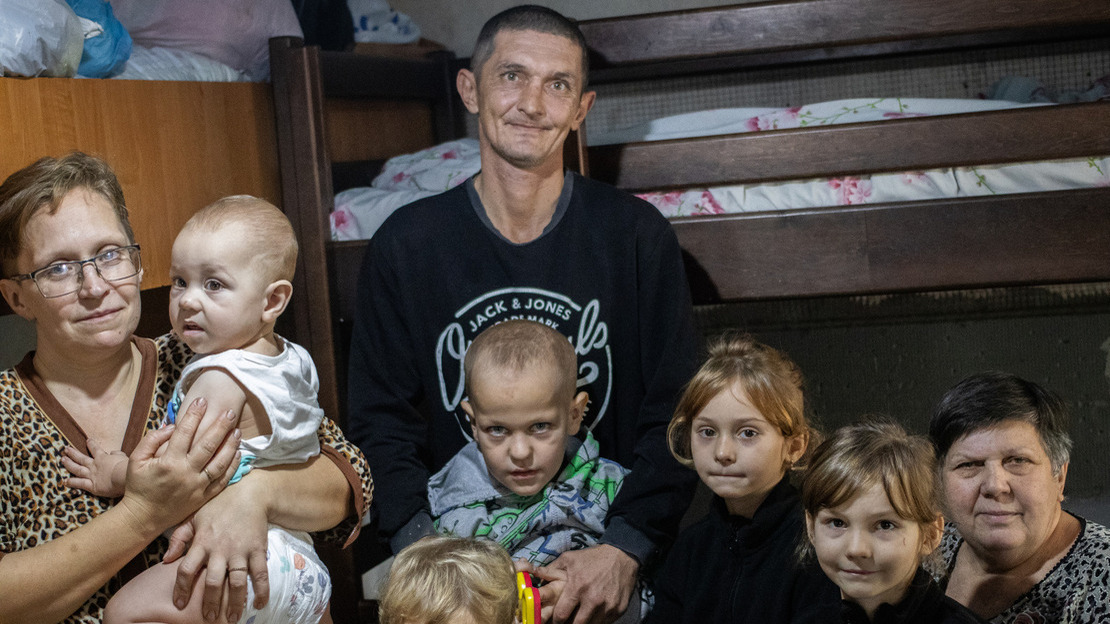 A large family is gathered in a room with bunk beds in the background. The father, in the center, has a gentle smile, while the mother, on the left, holds a baby in her arms. Around them, several children and their grandmother sit or stand, looking at the camera with shy smiles. ; }}