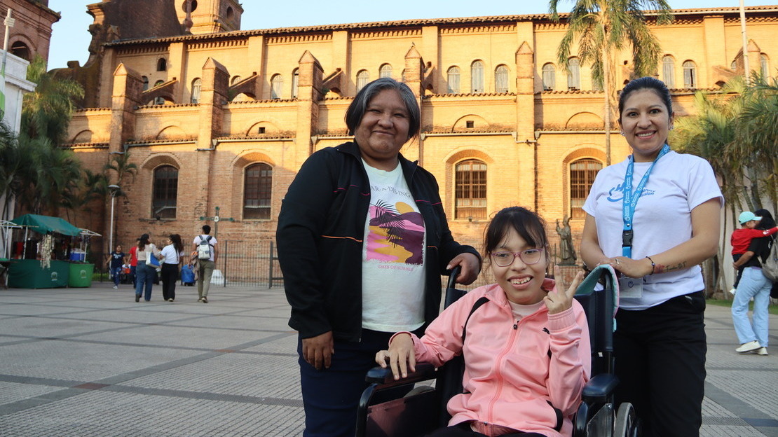 Trois femmes se tiennent sur une place, devant une cathédrale illuminée par le soleil. Deux d'entre elles sont debout et entourent la troisième, assises sur un fauteuil roulant. Elles sourient à la caméra.; }}