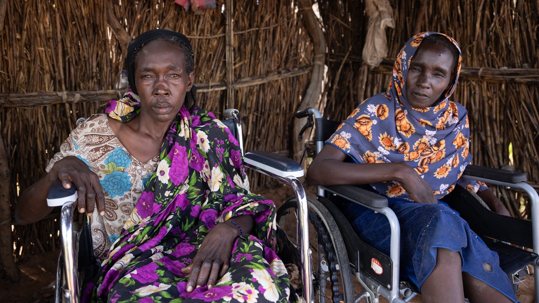 Kaltouma et Hassaneih sont côte à côte, chacun dans son fauteuil roulant. Elles sont abrités dans une maison en bois, et regardent l'objectif. Katlouma, à gauche, porte une robe avec un tissu à fleurs violet et blanc, tandis qu'Hassaneih, à droite, porte un foulard bleu à fleurs oranges. ; }}