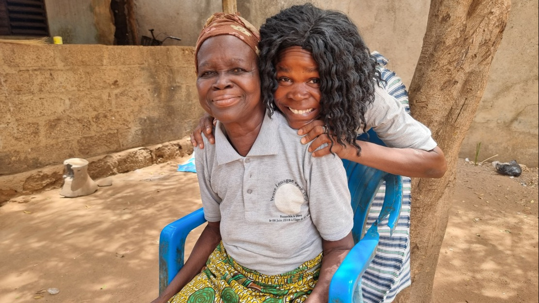 Likoulenan, a user of the African Network for Well-being and Resilience, and her mother. Savanes region, Togo. A young woman embraces an older woman sitting on a chair in a courtyard made of rammed earth. They both smile at the camera.; }}