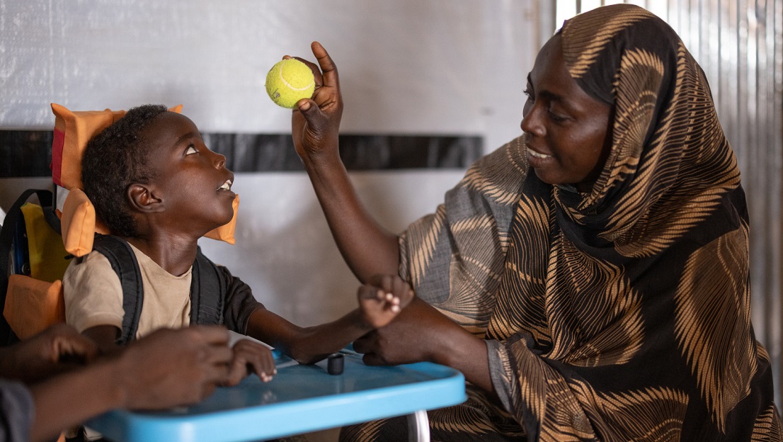 La photo est en intérieur, dans une salle de réadaptation d'HI. Omran, à gauche, est dans un fauteuil adapté et regarde la maman qui lui montre une balle de tennis. Djimilla, sa maman, regarde Omran en souriant. ; }}