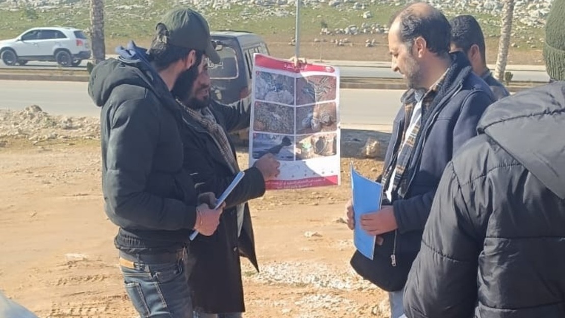 Teams show two men a poster with different examples of unexploded ordnance found in Syria. ; }}