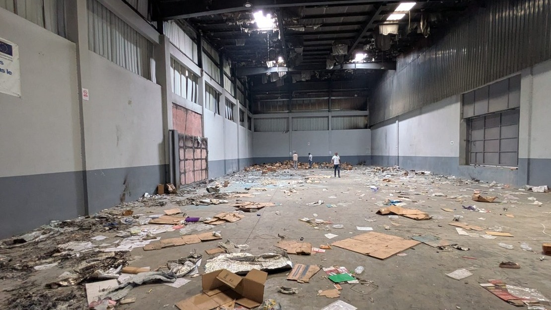 Inside a warehouse: the floor is littered with cardboard boxes, sacks and garbage of all kinds. Part of the roof is black with soot, and sheet metal panels are peeling off.; }}