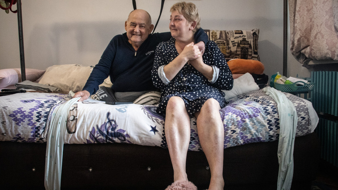 An elderly couple sits on a bed, sharing a moment of closeness. The man, dressed in a dark sweater, smiles warmly, while the woman, wearing a star-patterned robe and pink slippers, gently holds his hand over his shoulder. The intimate atmosphere of the room, with its colorful bedding and everyday objects, creates a feeling of warmth and connection.; }}